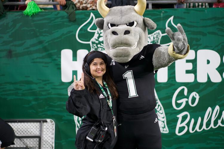 Risa Slepikas wearing an event headset at a USF Bulls game alongside the Rocky D. Bull mascot on the stadium sideline